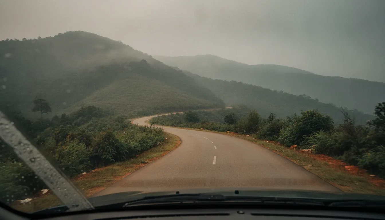 View through a rain-speckled car windshield of a winding road curving through misty green hills on the drive from Bangalore to Sakleshpur